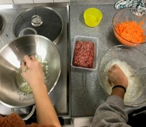 Personen bereiten Zutaten in einer Küche vor, darunter gehackte Zwiebeln, geriebene Karotten und rohes Hackfleisch.