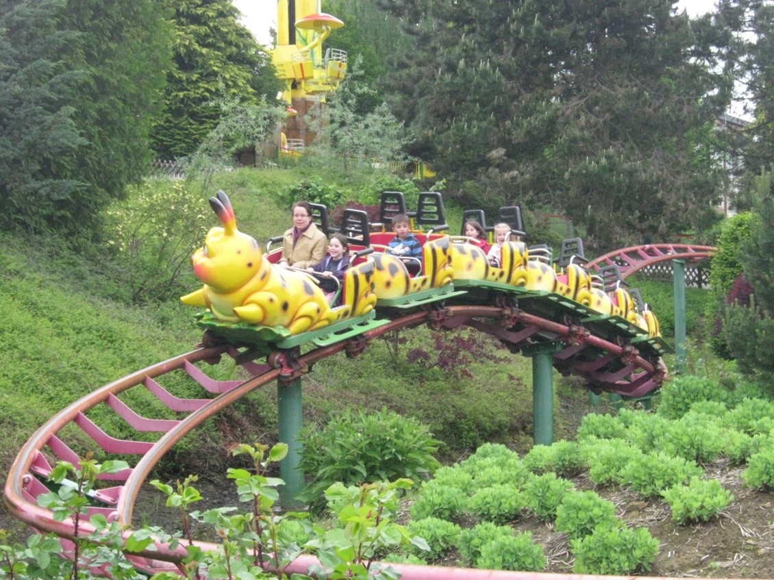 Eine bunte Raupen-Achterbahn fährt durch eine grüne, bewachsene Landschaft in einem Freizeitpark. Mehrere Personen sitzen in den Wagen und genießen die Fahrt.