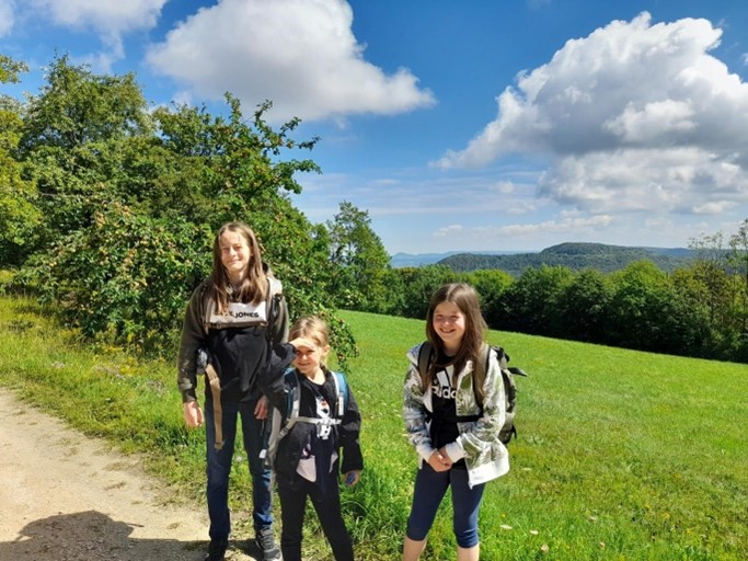 Drei Kinder stehen auf einem Wanderweg neben einer grünen Wiese und einem Wald, im Hintergrund sind Hügel und ein bewölkter Himmel zu sehen. Alle tragen Rucksäcke und sind für eine Wanderung gekleidet.