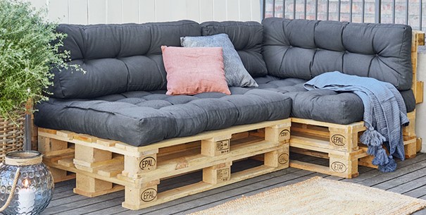 Gemütliches Sofa aus Europaletten mit grauen Polstern, dekoriert mit einem rosa und einem blauen Kissen sowie einer blauen Decke, auf einer Terrasse neben einer Pflanze und einer Laterne.