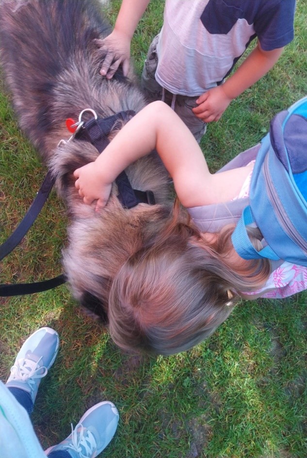 Zwei kleine Kinder streicheln und umarmen einen großen Hund im Freien. Das Mädchen trägt einen Rucksack und das Bild zeigt auch teilweise die Füße eines Erwachsenen.