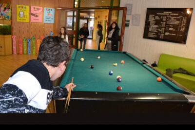 Ein Junge spielt Billard in einem Freizeitraum, während andere Personen im Hintergrund stehen. An der Wand hängen bunte Schilder mit verschiedenen Aufschriften.