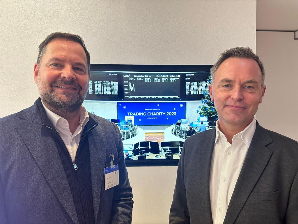Zwei Männer stehen vor einem Bildschirm, der Informationen zum Trading Charity 2023 anzeigt.