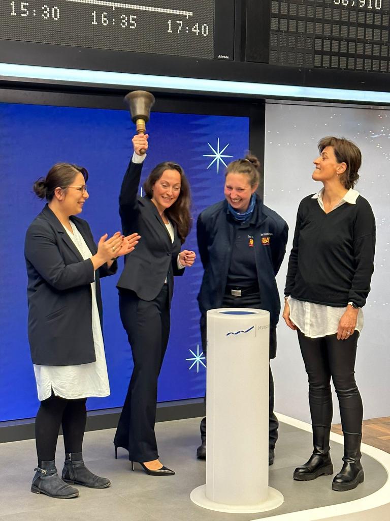 Eine Gruppe von 4 Frauen steht vor einer digitalen Anzeige und feiert, wobei eine Person eine Glocke hochhält.