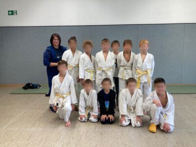 Eine Gruppe von Kindern in weißen Judo-Anzügen posiert für ein Foto in einer Sporthalle. Die Kinder tragen gelbe Gürtel und knien oder stehen auf einem hellen Boden vor einer grauen Wand. Links im Bild ist eine Person in einem blauen Trainingsanzug zu sehen.