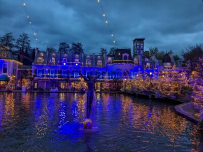 Beleuchtete Gebäude und Bäume am Ufer eines Gewässers in einem Freizeitpark bei Dämmerung, mit einer Statue im Vordergrund und Lichterketten, die über den Himmel gespannt sind.