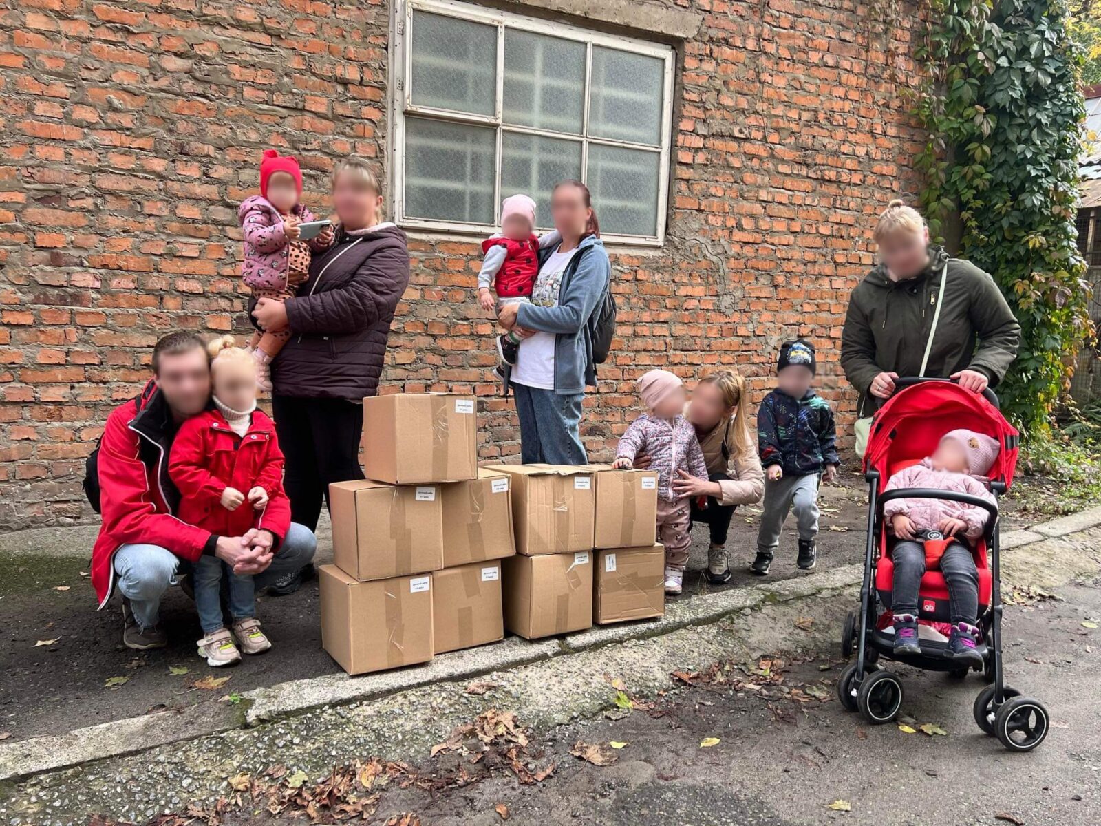 Eine Gruppe von Menschen, darunter mehrere Kinder, steht vor einer Backsteinwand und neben gestapelten Kartons. Ein Kind sitzt in einem roten Kinderwagen.
