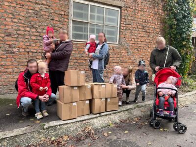 Eine Gruppe von Menschen, darunter mehrere Kinder, steht vor einer Backsteinwand und neben gestapelten Kartons. Ein Kind sitzt in einem roten Kinderwagen.