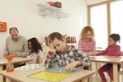 In einem Klassenraum sitzen einige Kinder und versuchen Aufgaben zu lösen. Neben jedem Kind sitzt eine Erwachsene Person die bei der Lösung der Aufgaben unterstützt.