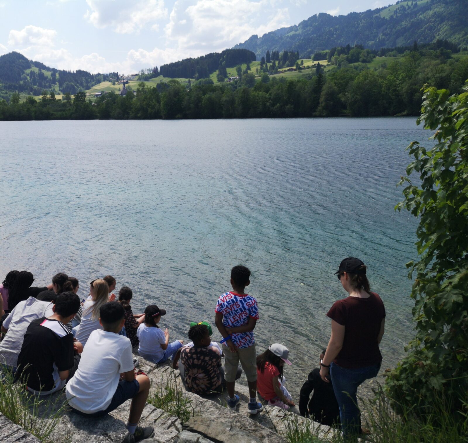 Ein See umgeben von Bäumen. In der Mitte sitzen einige Kinder. Man sieht sie nur von Hinten wie sie auf das Wasser schauen.