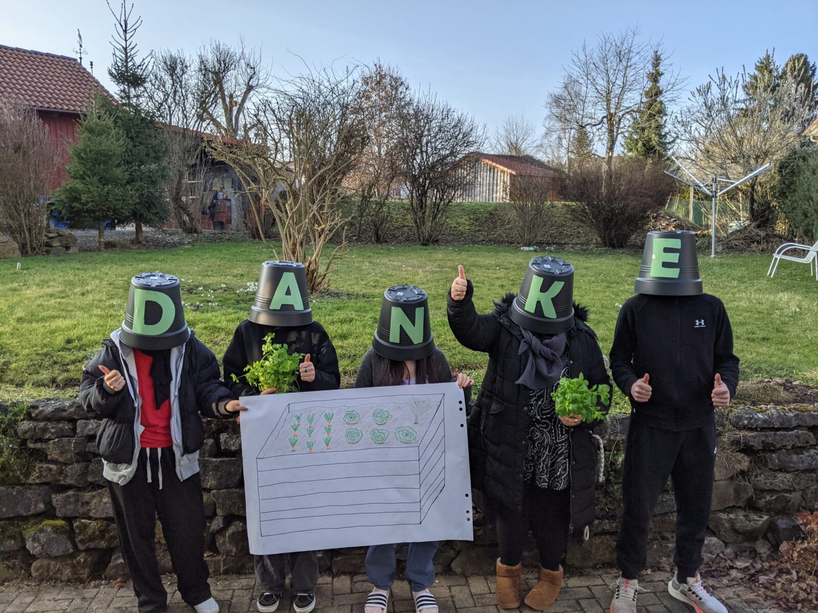 Fünf Personen stehen draußen vor einer Steinmauer und halten ein Schild mit einer Zeichnung eines Hochbeets. Sie tragen Eimer auf dem Kopf, die jeweils einen Buchstaben des Wortes "DANKE" bilden. Einige Personen halten Pflanzen in den Händen und zeigen Daumen hoch. Im Hintergrund ist ein Garten mit Bäumen und Sträuchern zu sehen.