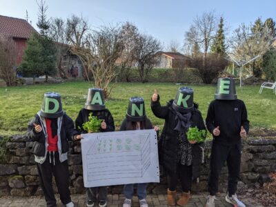 Fünf Personen stehen draußen vor einer Steinmauer und halten ein Schild mit einer Zeichnung eines Hochbeets. Sie tragen Eimer auf dem Kopf, die jeweils einen Buchstaben des Wortes "DANKE" bilden. Einige Personen halten Pflanzen in den Händen und zeigen Daumen hoch. Im Hintergrund ist ein Garten mit Bäumen und Sträuchern zu sehen.