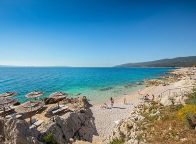 Ein sonniger Strand mit klarem, türkisfarbenem Wasser und felsiger Küste. Mehrere Strohdach-Sonnenschirme und Liegestühle stehen auf den Felsen. Menschen genießen den Strand und das Wasser. Im Hintergrund sind bewaldete Hügel und eine weite Meereslandschaft zu sehen.