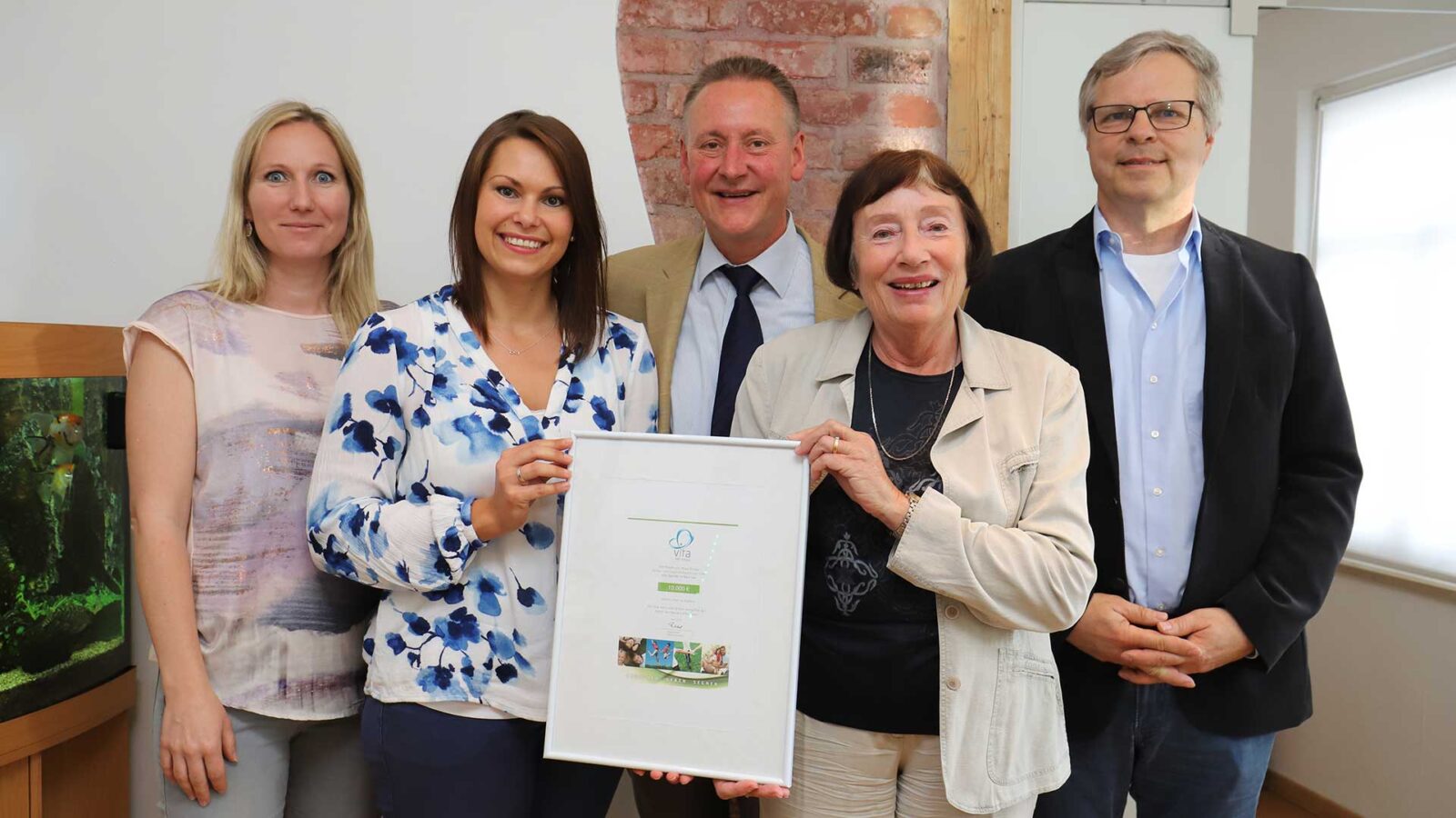 Eine Gruppe von Erwachsenen steht vor einer Wand und hält eine gerahmte Urkunde. Im Hintergrund ist ein Aquarium zu sehen.