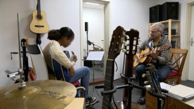 Zwei Personen - ein Mädchen und ein Mann - spielen Akustikgitarren in einem Musikraum, umgeben von verschiedenen Musikinstrumenten und Notenständern.