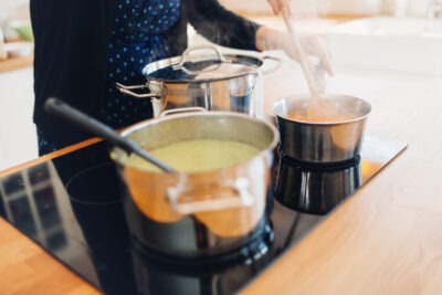 Person kocht auf einem modernen Induktionsherd, zwei Töpfe mit dampfenden Speisen, eine grüne Suppe und eine rote Soße werden gerührt.