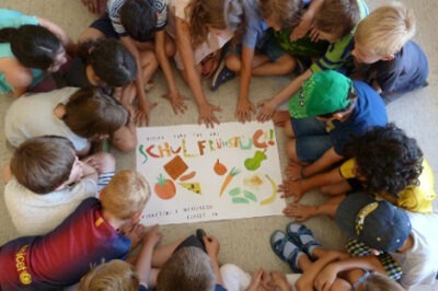 Kinder sitzen im Kreis und betrachten ein Poster mit der Aufschrift "Schulfrühstück" und Bildern von Lebensmitteln.
