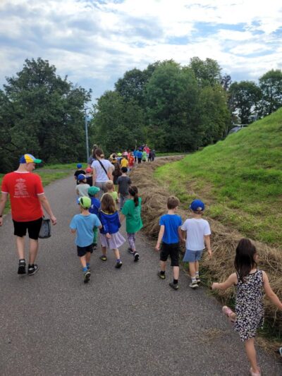 Eine Gruppe von Kindern und Erwachsenen spaziert auf einem gepflasterten Weg durch eine grüne Landschaft. Die Kinder tragen bunte Kleidung und einige von ihnen haben Hüte auf. Der Himmel ist bewölkt.