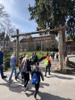 Eine Gruppe Kinder und Erwachsener laufen gemeinsam einen Weg entlang. Auf dem Weg ist aus Holzpfälern ein Durchgang gebaut wurden mit einem Schild "SOMMERBERG".