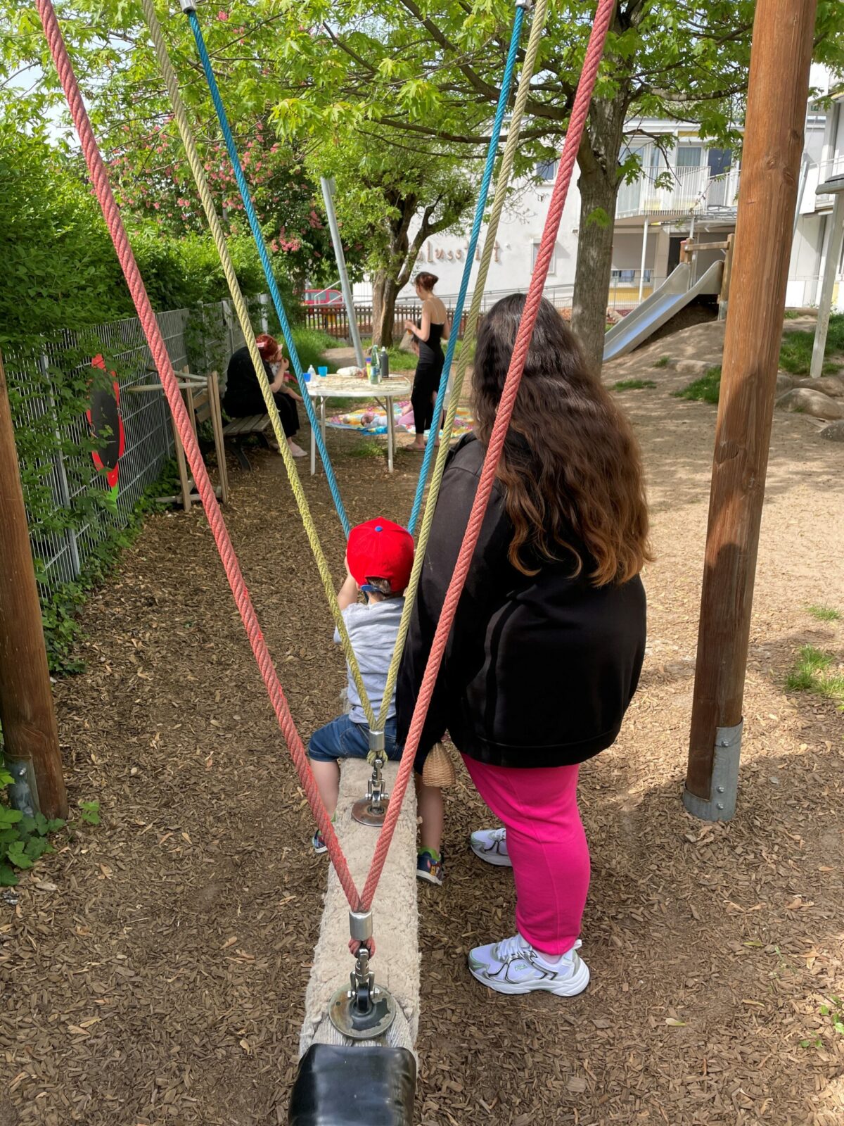 Auf dem Bild ist eine Frau mit einem Kind zu sehen, die auf einem Spielplatz sind. Das Kind sitzt auf einer Schaukel und die Frau hält das Kind am Rücken fest.