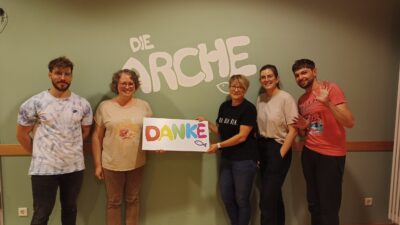 5 Erwachsene stehen vor einer Grünen Wand. Auf der grünen Wand steht "DIE ARCHE". Die zwei Erwachsenen in der Mitte halten ein Plakat mit der Aufschrift "DANKE". Sie schauen alle lächelnd und freundlich in die Kamera.