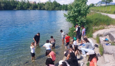 Eine Gruppe von Menschen steht am Ufer eines Sees, einige von ihnen mit den Füßen im Wasser. Sie genießen die Natur und unterhalten sich.