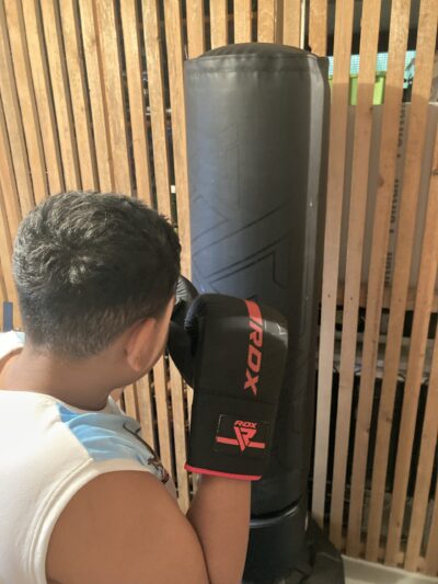 Ein Junge mit kurzen Haaren trägt Boxhandschuhe und trainiert an einem schwarzen Boxsack in einem Raum mit Holzwänden.