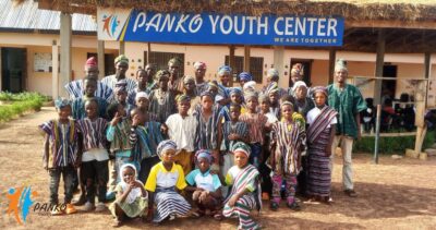 Eine große Gruppe von Kindern und Erwachsenen posiert vor einem Gebäude mit der Aufschrift „PANKO Youth Center“. Sie tragen bunte Kleidung und stehen dicht beieinander.