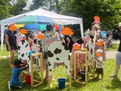 Kinder spielen unter einem weißen Pavillon mit Holzfiguren, die Kühen nachempfunden sind. Sie tragen bunte Kleidung und es handelt sich hierbei um ein Sommerfest.