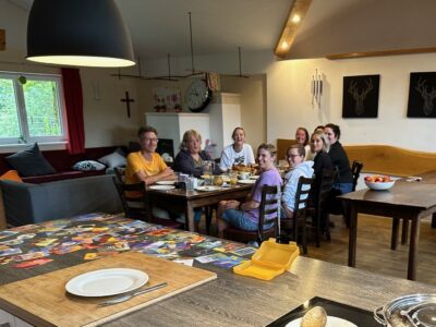 Mehrere Erwachsene sitzen an einem langen Tisch in einem gemütlichen Raum und unterhalten sich. Der Tisch ist gedeckt mit Tellern, Tassen und einem Frühstücksbuffet.