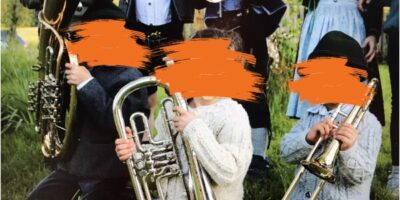 Mehrere Musiker in traditioneller Tracht spielen verschiedene Blasinstrumente, darunter eine Tuba und eine Trompete. Die Szene spielt sich im Freien ab, mit Bäumen im Hintergrund.