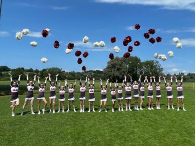 Eine reihe junger Mädels steht nebeneinander in ihren Cheer-Outfits und werfen ihre Pompons in die Höhe.