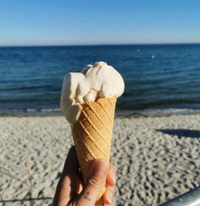 Eine Hand hält eine Eiswaffel mit zwei Kugeln Vanilleeis vor einem traumhaften Strand. Das Meer ist ruhig, und der Himmel zeigt sich leicht bewölkt, aber dennoch sonnig. Die Aufnahme vermittelt ein Gefühl von Sommer, Entspannung und Genuss.