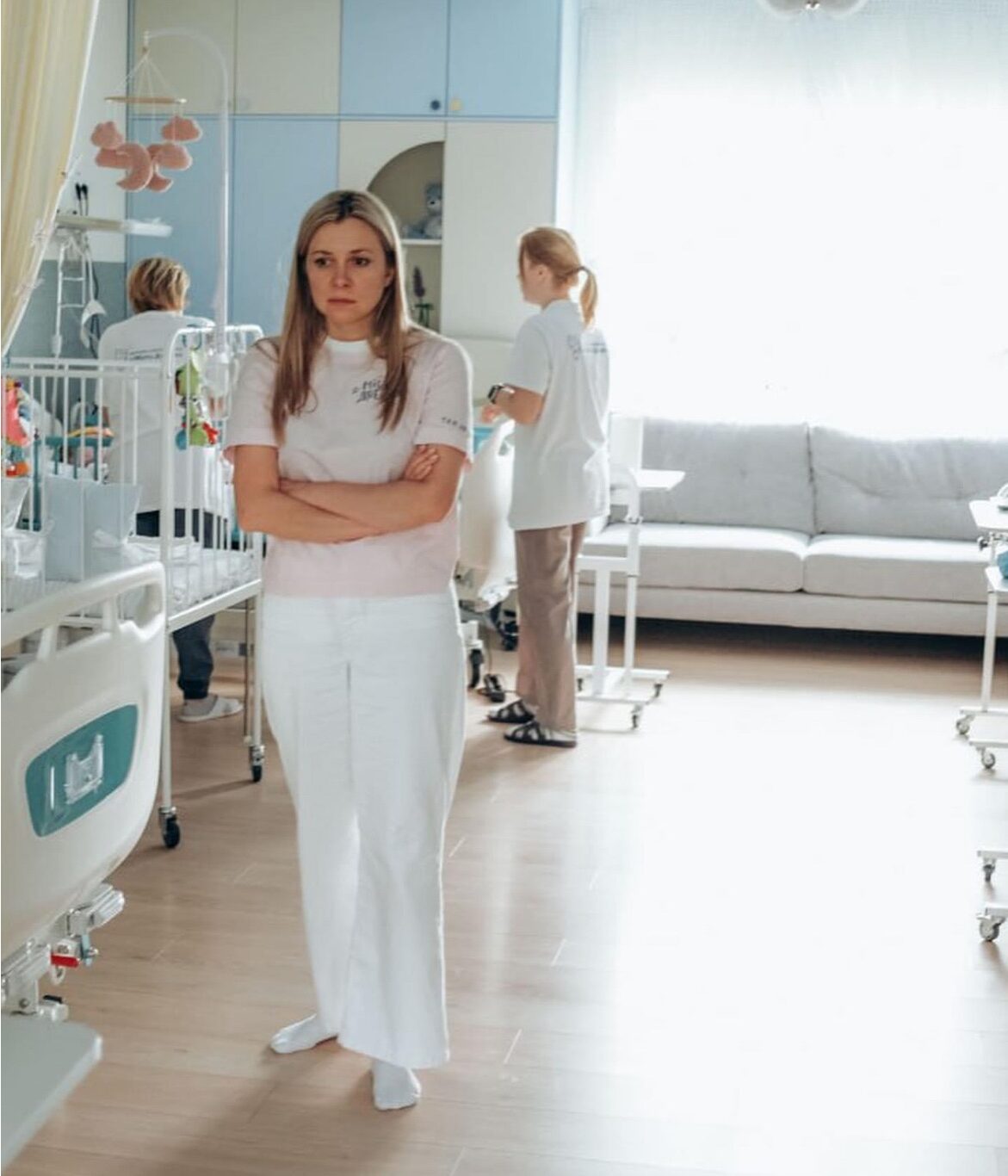 Eine Frau in weißer Kleidung steht mit verschränkten Armen in einem Krankenhausflur. Hinter ihr befinden sich Kinderbetten und ein heller Aufenthaltsraum.
