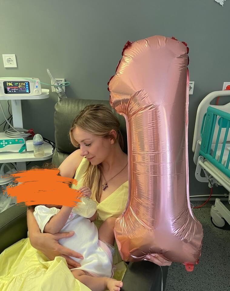 Das Bild zeigt eine junge Frau in einem Krankenzimmer. Sie sitzt auf einem Stuhl und hält in der einen Hand einen Helliumluftbalon in der Zahl 1 und mit der anderen Hand hält sie ein Baby. Die Frau lächelt das Baby freundlich an.