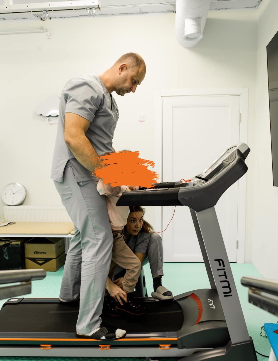 Ein Mann in Sportkleidung unterstützt ein Kind, das auf einem Laufband steht. Die Szene spielt sich in einem Rehabilitationszentrum oder einer medizinischen Einrichtung ab.