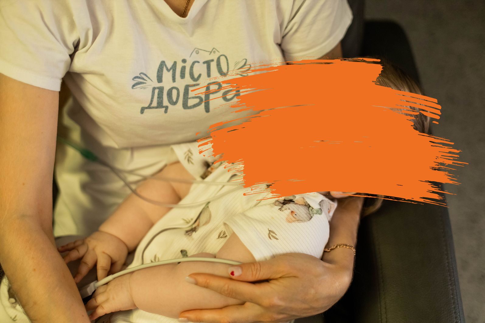Eine Frau trägt ein Shirt mit der Aufschrift „Misto Dobra“, sitzt auf einem Sessel und hält ein Baby in den Armen.