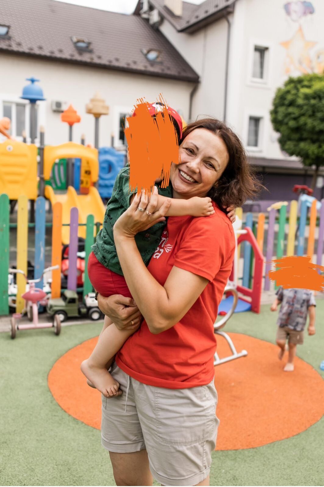 Eine fröhliche Frau in einem roten Shirt hält ein kleines Kind auf dem Arm, das ebenfalls lacht. Sie stehen auf einem bunten Spielplatz mit vielen Spielgeräten.