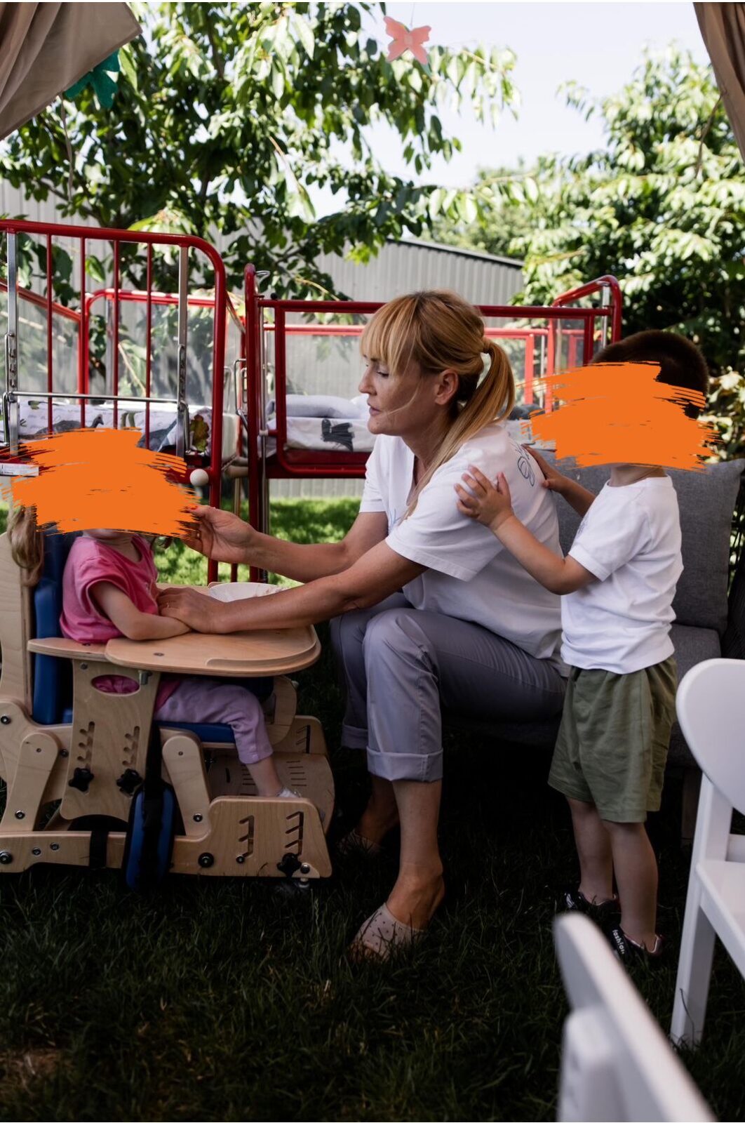 Eine Frau betreut zwei kleine Kinder auf einem Spielplatz. Sie kniet sich zu ihnen hinunter und interagiert mit ihnen an einem bunten Spielgerät
