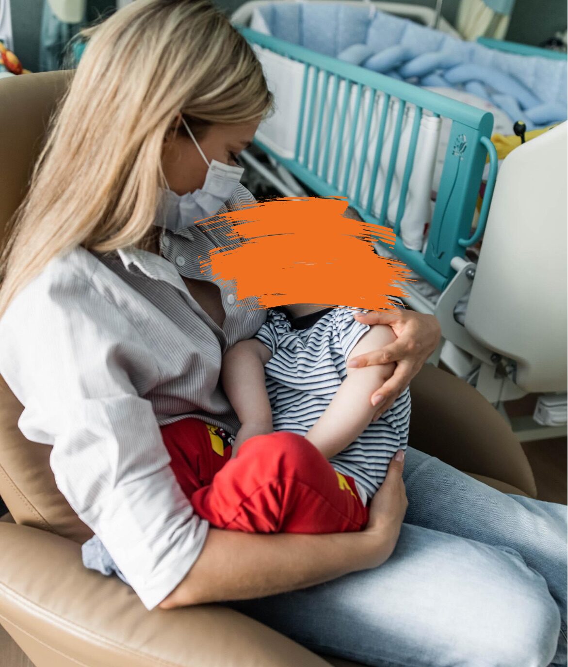 Eine Frau mit Schutzmaske hält ihr Kind, das eine rote Hose und ein blau-weiß gestreiftes Oberteil trägt. Sie sitzt auf einem Stuhl neben einem Krankenhausbett.