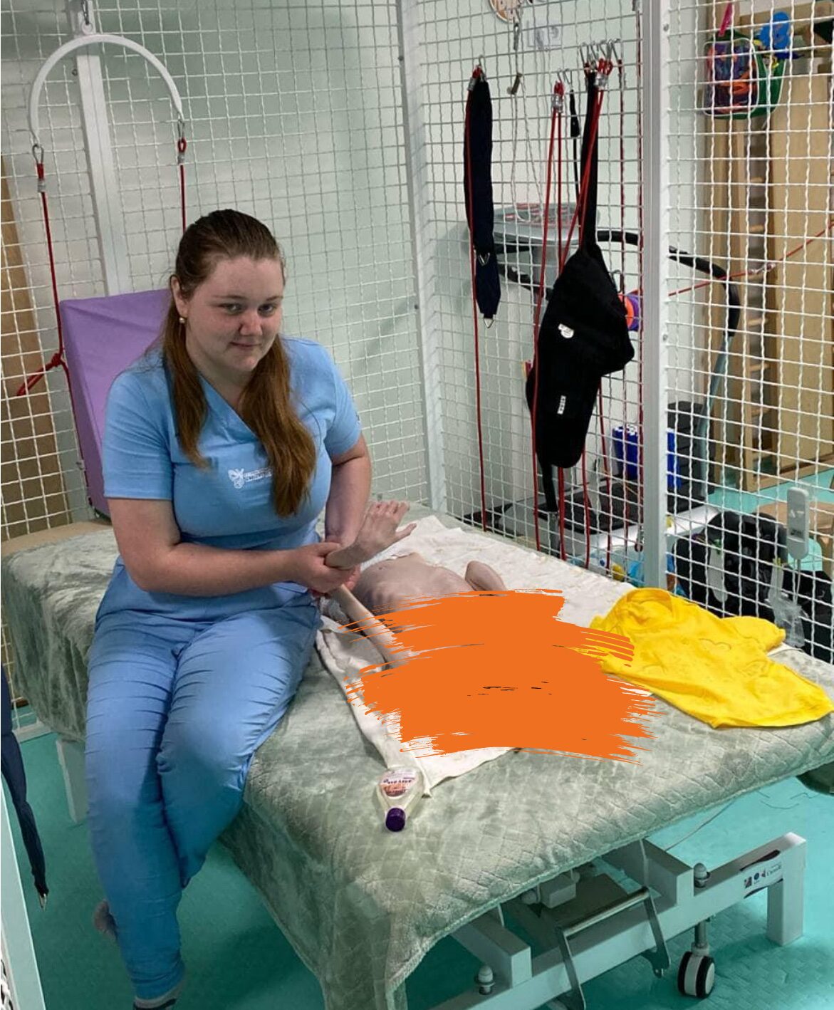 Eine junge Frau in blauer Berufskleidung sitzt neben einem Kind auf einer Behandlungsliege in einer Reha-Einrichtung. Sie scheint therapeutische Übungen mit dem Kind durchzuführen.