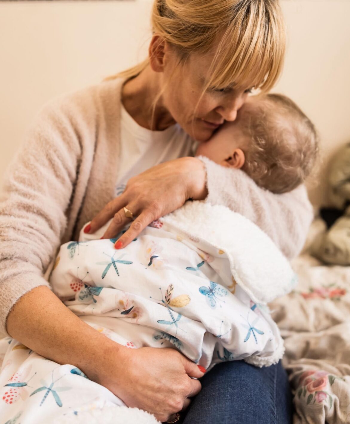 Eine Frau sitzt mit einem Baby auf dem Arm und hält es liebevoll an sich gedrückt. Sie scheint Trost zu spenden, während das Baby in eine Decke eingewickelt ist.
