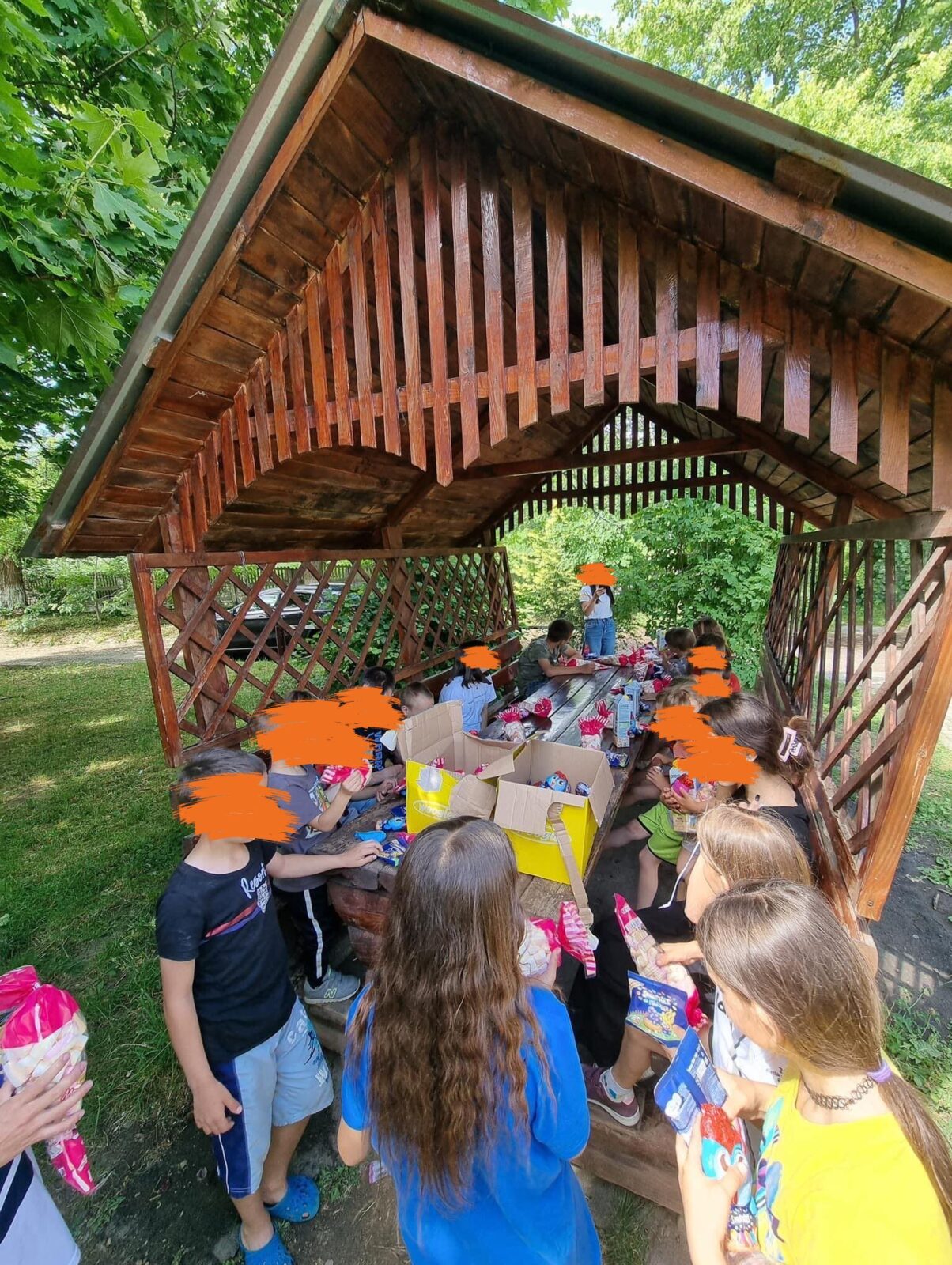 Eine große Gruppe von Kindern sitzt unter einem überdachten Holzpavillon im Freien. Sie scheinen an einer Veranstaltung oder einem Workshop teilzunehmen. Im Hintergrund sind Bäume zu sehen.