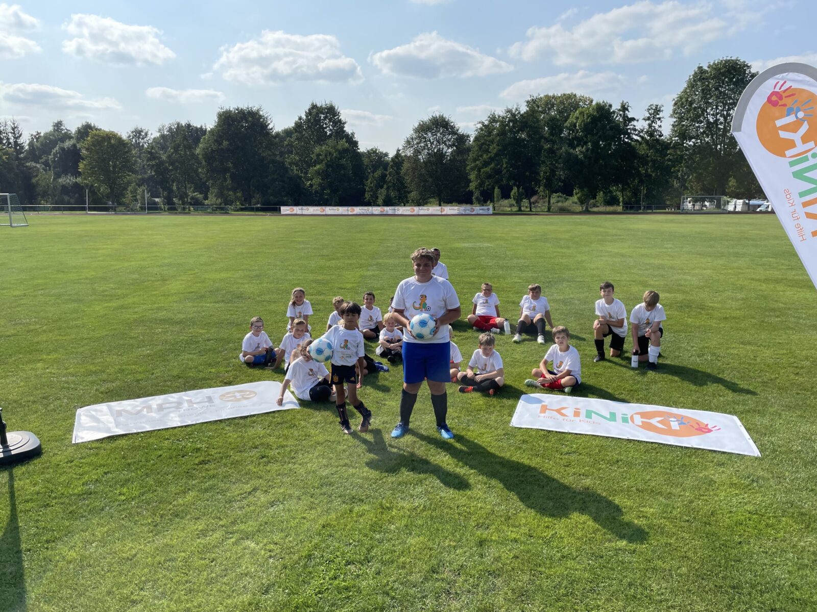 Eine Gruppe Kinder sitzen in weißen T-Shirts auf einer Wiese. Vor ihnen liegen zwei Banner. Auf dem einen Banner rechts ist das KiNiKi Logo drauf und auf dem Banner links ist das MRH Trowe Logo drauf. Eine Frau steht vor den Kindern und hält einen Fußball in der Hand.