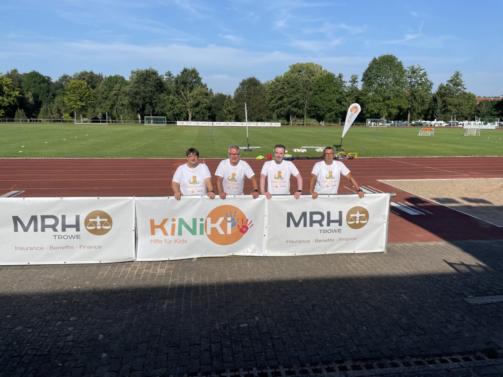 Ein großes weißes Banner mit den Logos von „KiNiKi“ und „MRH Trowe“ ist an einem Zaun befestigt. Hinter dem Banner stehen mehrere Erwachsene in weißen T-Shirts. Der Rasen im Hintergrund ist gepflegt und es sind weitere Teilnehmer sowie Sporteinrichtungen zu sehen.