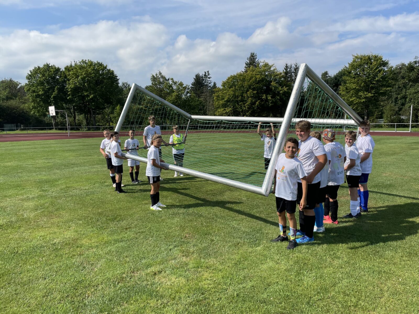 Mehrere Erwachsene und Kinder in weißen T-Shirts sind auf einem großen Sportfeld zu sehen. Sie tragen gemeinsam ein großes Fußballtor und scheinen es an eine neue Position zu bringen.