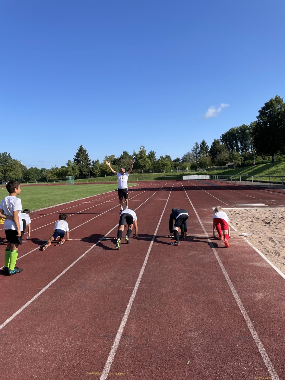 Mehrere Kinder stehen in Startposition auf einer roten Laufbahn, bereit für ein Wettrennen. Ein Erwachsener steht im Hintergrund und gibt ihnen das Startsignal. Die Laufbahn liegt auf einem Sportplatz, umgeben von einer grünen Wiese und Bäumen. Die Kinder tragen sportliche Kleidung, einige haben ihre Hände auf dem Boden, während sie sich auf den Start konzentrieren.