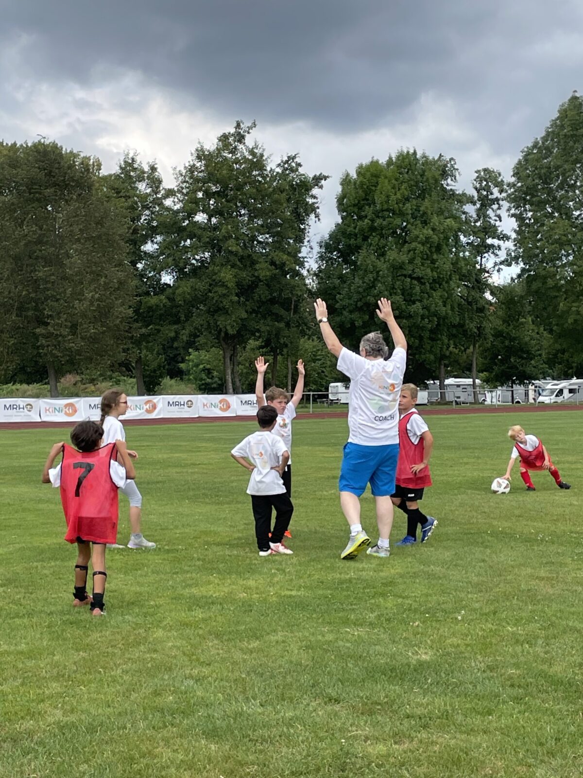 Kinder spielen Fußball auf einer grünen Wiese unter bewölktem Himmel. Ein Trainer in blauer Shorts und weißen Socken hebt die Arme, während mehrere Kinder in weißen und roten Trikots ebenfalls ihre Hände heben. Im Hintergrund sind Bäume und eine Laufbahn zu sehen.