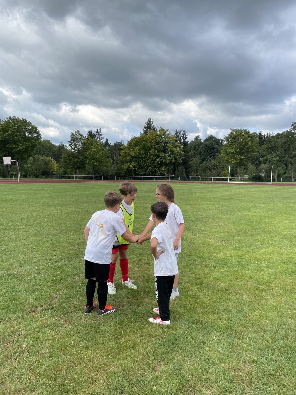 4 Jungen stehen in Fußballklamotten auf einer Wiese. Sie stehen im Kreis und haben jeweils einen Arm in die Mitte gestreckt und die Hände übereinander gelegt. Im Hintergrund sind Bäume und ein grauer Himmel zu sehen.