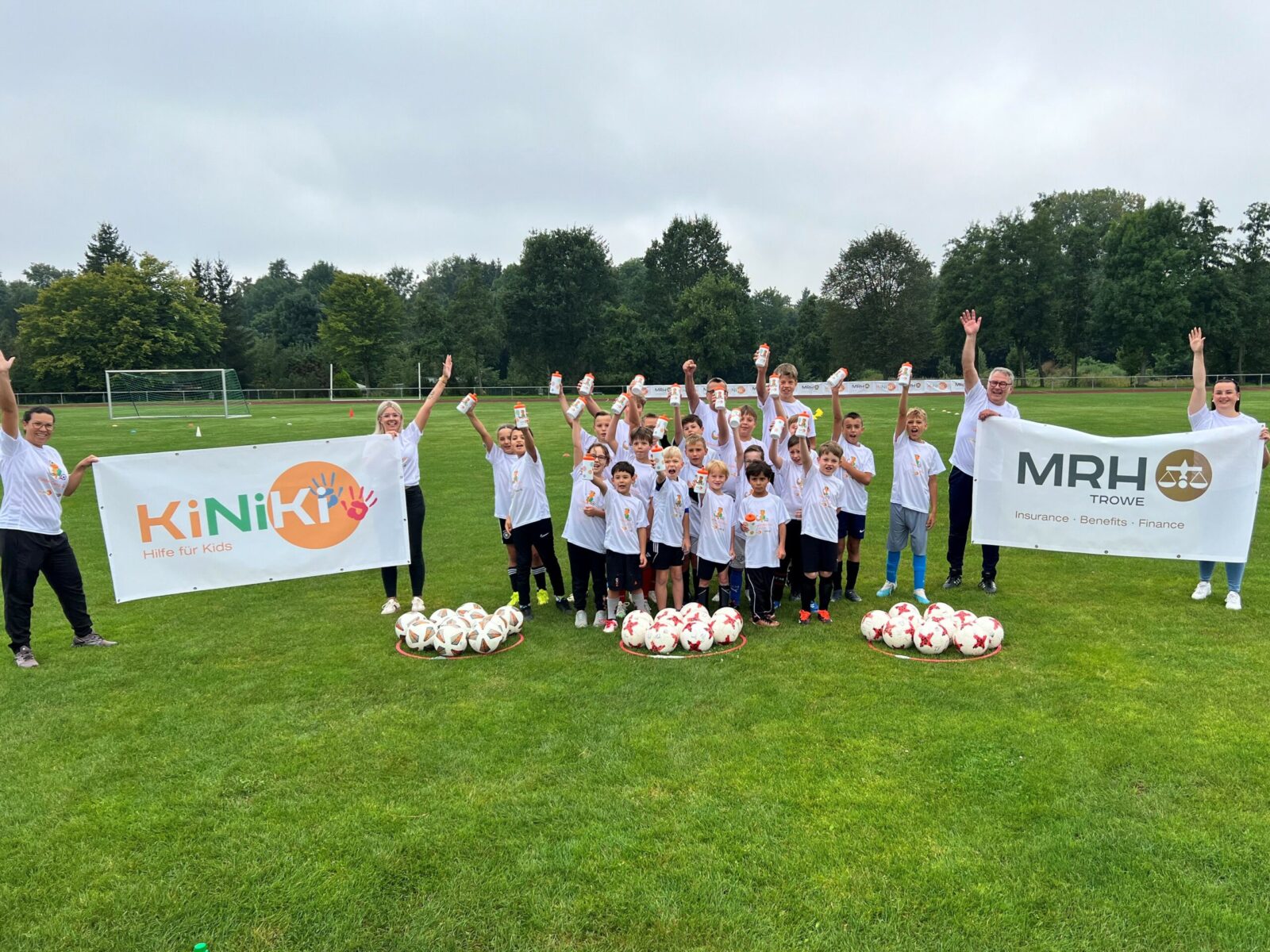 Eine Gruppe von Kindern steht auf einem Fußballfeld, vor sich mehrere weiße Fußbälle aufgereiht. Mit erhobenen Armen jubeln sie in die Kamera, während sie breit lächeln. Rechts und links neben der Gruppe stehen jeweils zwei Erwachsene, die je gemeinsam einen Banner hochhalten. Auf dem Banner rechts ist das MRH Trowe Logo drauf und auf dem Banner links das KiNiKi Logo.